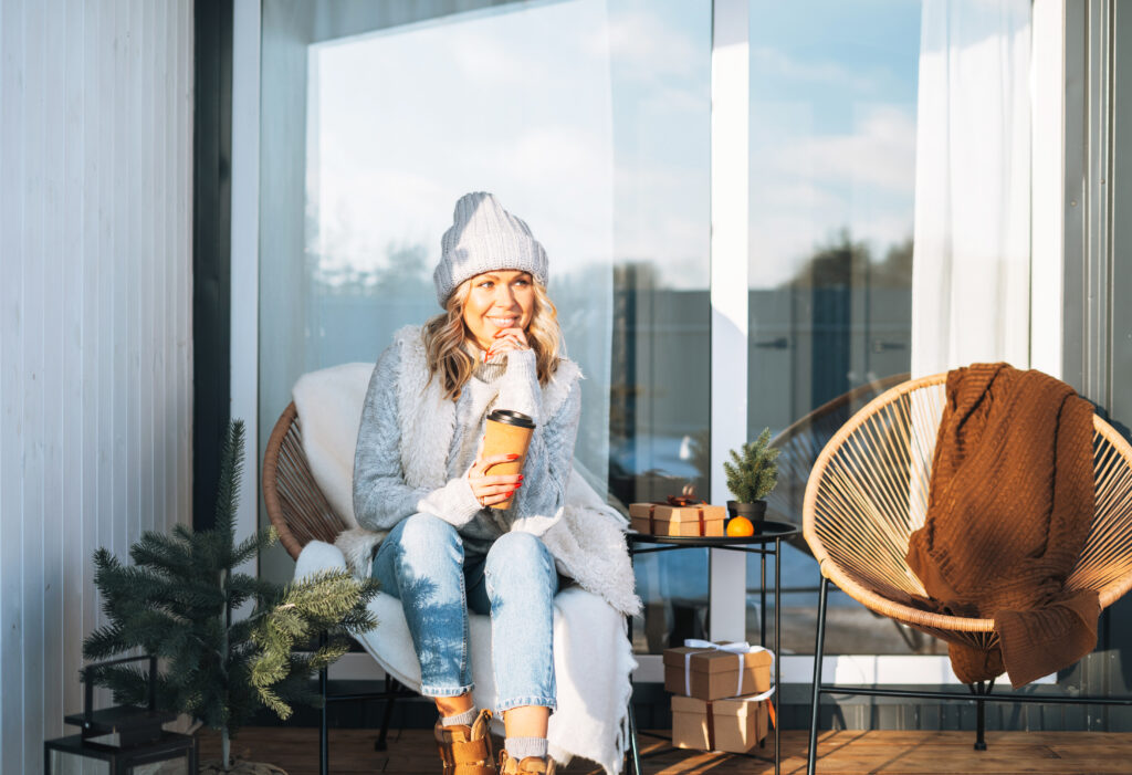 A woman with a scarf and a hat sitting on the porch drinking coffee.