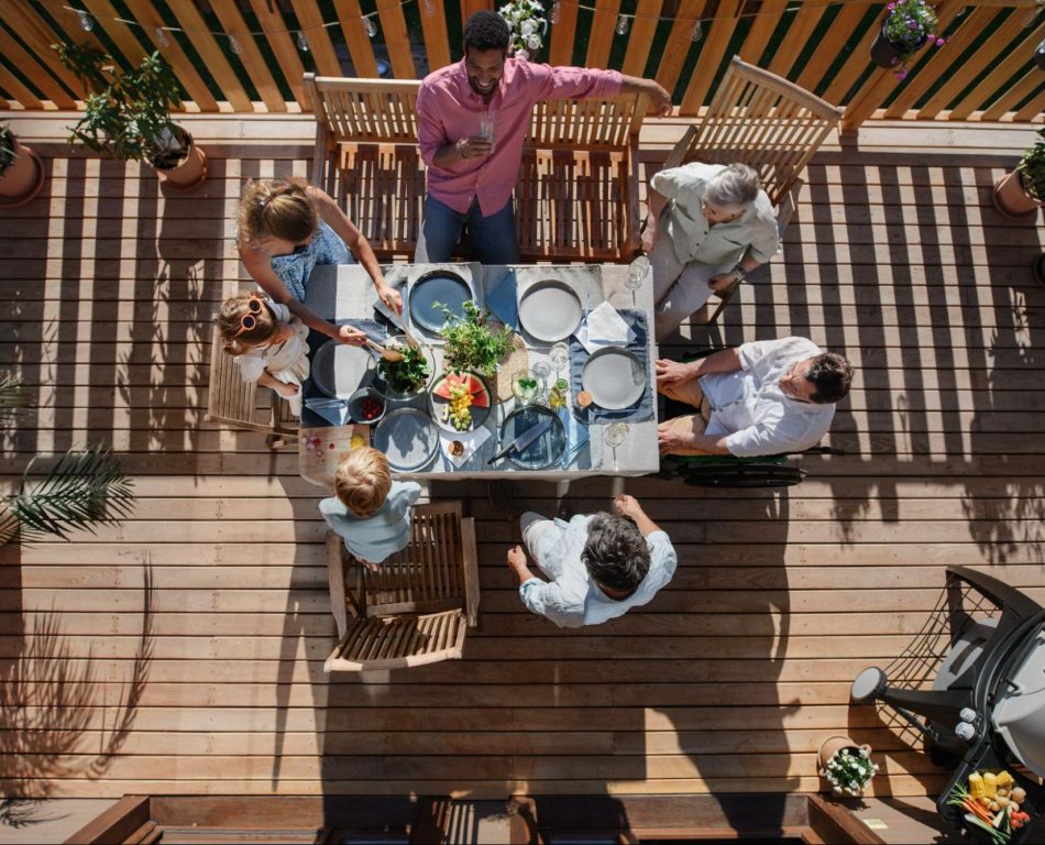 Group of people sitting around a dining table on a deck made of wood with plants and a grill in the background.